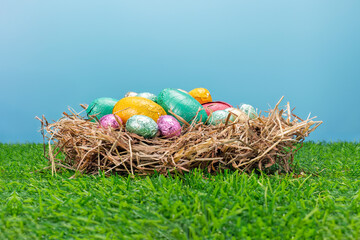 Colorful Foil Wrapped Chocolate Easter Eggs in Straw Nest on Green Artificial Grass with Blue Sky Background