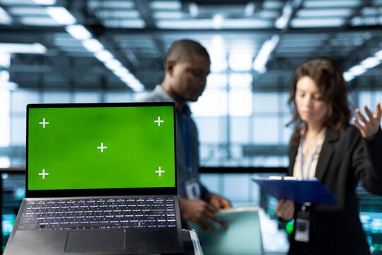 Green screen laptop in front of data center technicians in blurry background calibrating energy usage, inspecting UPS configurations, supporting sustainable infrastructure