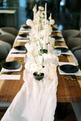 Festive table with bouquets of white flowers on a narrow tablecloth, candles and black crockery