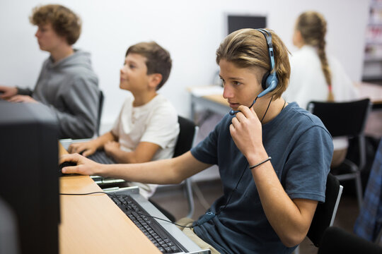 Portrait of a concentrated fifteen-year-old schoolboy studying at a computer wearing headphones in class at lesson