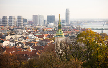 Scenic autumn view of Bratislava with medieval St. Martin Cathedral rising above historic terracotta tiled rooftops against backdrop of modern skyscrapers on bank of Danube River, Slovakia