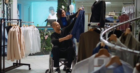 Asian woman helping wheelchair user choose the best clothing items at a mall store, browsing for the new collection of designer merchandise. Diverse customers on a shopping spree. Camera B.