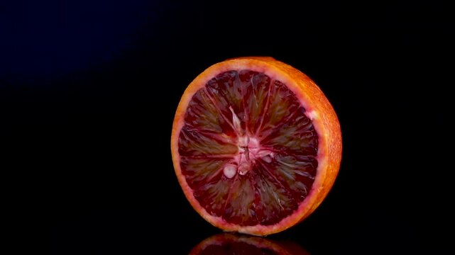 A juicy, sliced Sicilian red orange rotating on a black background with drops of fresh juice dripping down, showcasing its vibrant color and refreshing texture.