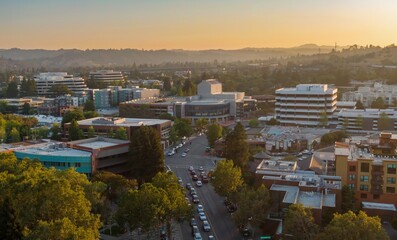 Aerial view of Walnut Creek, California, USA, at sunset. Cars drive on the road, surrounded by buildings and trees. The sun sets over the hills in the distance.