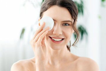 Joyful young lady removing makeup from her eyes, covering one eye with cotton pad and smiling at...