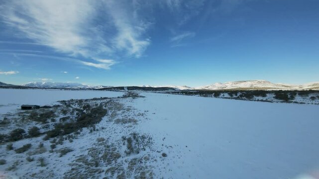 Aerial-FPV-First person view - Flying over snow covered field in shade of sun gone down behind the west mountain with sunlight still hitting distant hills 