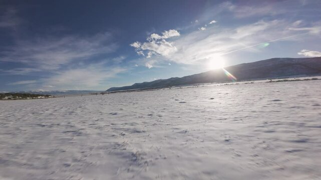 Aerial-First person view-FPV-Fast and low over snowy field and passing over irrigation wheel line-toward setting sun flare as last light of day lingers over the top of the mountain.