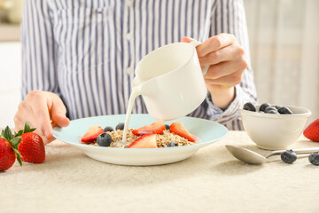 Woman pouring milk into bowl with oatmeal and berries at beige textured table, closeup