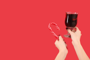 Female hands with glass of warm mulled wine and candy cane on red background