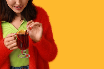 Beautiful young woman with glass cup of hot mulled wine on yellow background