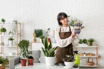 Beautiful young happy female gardener with houseplants wearing gardening gloves at home