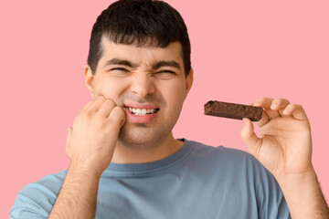 Young man with candy bar suffering from toothache on pink background, closeup