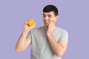 Young man with orange suffering from toothache on lilac background