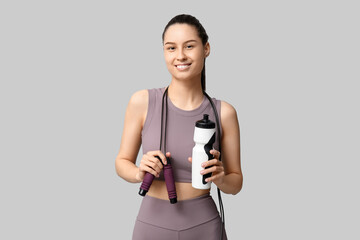 Young sporty woman sitting with water bottle and skipping rope on grey background
