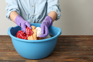 Woman in rubber gloves washing clothes in plastic basin on wooden table against light background