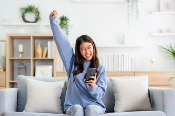 Happy young asian woman relax on comfortable couch at home texting messaging on smartphone, smiling...
