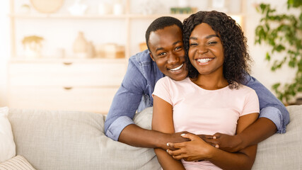 Obraz premium Cheerful african man hugging his smiling wife from behind, spending time together at home, empty space