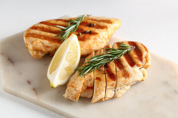 Board with tasty grilled chicken breast, rosemary, peppercorns and lemon slice on white background