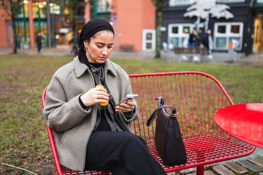 Student in niqab drinking coffee and using smartphone on city bench