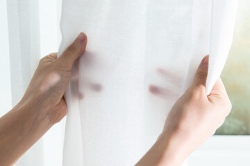 Close-up of woman hand opening white fabric curtain at home.