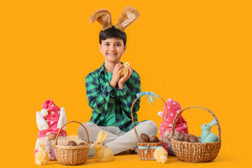 Cute boy in bunny ears with wicker baskets of sweet chocolate eggs and toys sitting on yellow background. Easter celebration