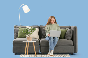 Beautiful young woman with laptop sitting on stylish grey sofa against blue background