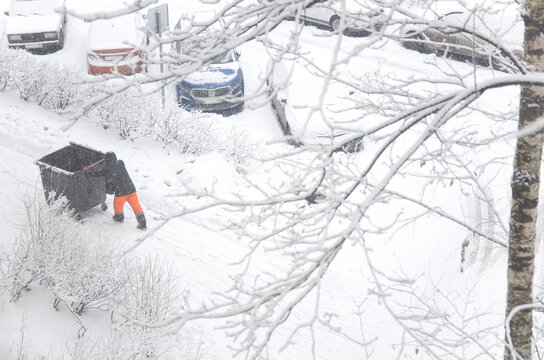 Snowfall blankets city street as worker hauls trash bin