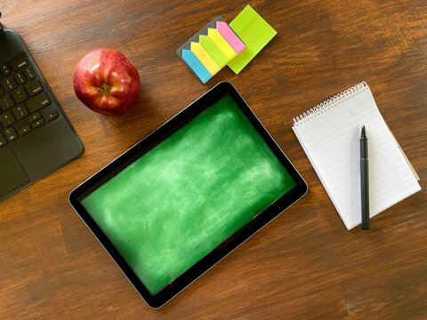 On a wooden table there is a tablet with a green background imitating a board, around it there is an apple, stickers and a notepad.
