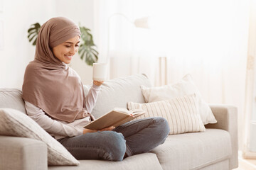 Fototapeta premium Weekend Pastime. Young Arab Woman In Hijab Relaxing With Book And Coffee On Couch, Enjoying Spending Time At Home, Copy Space