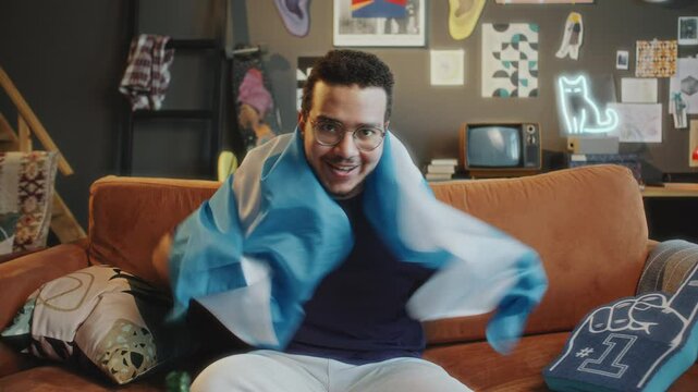 Excited sports fan sitting on couch in stylish living room, smiling and draping Argentine flag over shoulders while watching TV game and cheering for favorite team. Zoom shot