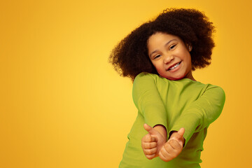 Everything is great. Positive cute little girl gesturing thumbs up and smiling at camera, yellow...