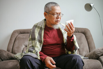 Sick Elderly Man Sneezing In Napkin Suffering From Fever And Flu While Sitting On Sofa In Living...
