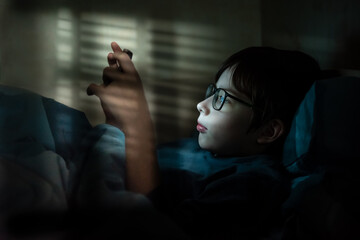 Teenage boy with glasses at night under a blanket with a smartphone. Children who are addicted to...