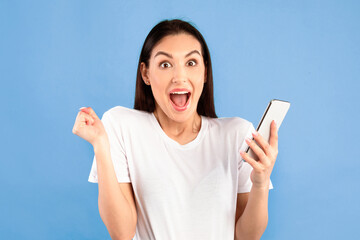 Yes. Portrait of happy surprised young woman using mobile phone making winner gesture isolated over blue studio wall background. Excited lady celebrating online win, screaming and looking at camera