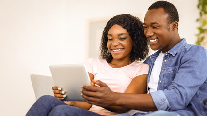 Fototapeta premium Closeup of black family husband and wife using digital tablet, sitting on couch at living room, empty space. Smiling african american man and woman holding brand new pad, using new exciting app