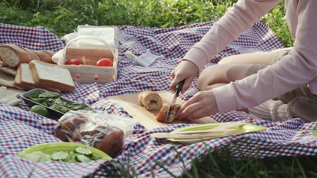 A teenage girl in a pink sweater kneels on a plaid blanket at a picnic and cuts a chocolate filled croissant with a kitchen knife on a sunny summer day. Medium shot