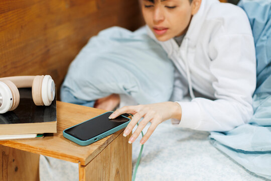 Young woman in cozy bedroom checking time in phone from bed