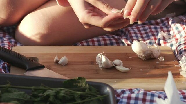 The hands of a teenage girl sitting on a plaid blanket at a picnic peel garlic for preparing sauce on a sunny summer day. Front view, close up shot