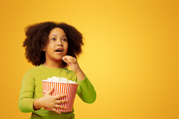 Movie Premiere. Excited cute little african girl eating popcorn and looking away at something with...