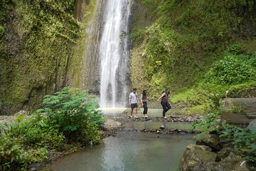 group of young asian indonesian man and women walking and crossing small stream with waterfall on the background