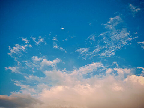 Bright blue sky with wispy white clouds and a small moon. Lower clouds have warm reddish tones suggesting sunrise or sunset.