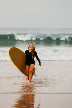 A smiling female surfer walks back from the ocean carrying her b