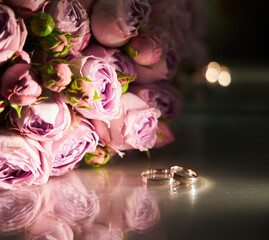 Two wedding rings are on a table next to a bouquet of pink flowers. The rings are shiny and reflect the light, creating a romantic and elegant atmosphere