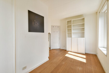 A bright and minimalist room featuring light hardwood flooring, a built-in shelf, and large windows letting in sunlight, creating a serene atmosphere.