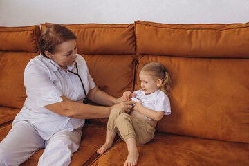 Doctor woman doing examination to little girl child baby sitting on sofa, home care