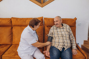 Female doctor measures blood pressure of elderly man pensioner sitting on sofa, home care