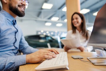 Close up of hands typing on keyboard at car salon with customer smiling at contract in background.