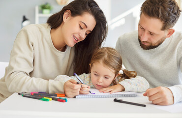 Little cute child girl sitting at the desk with young parents in living room at home and drawing a...