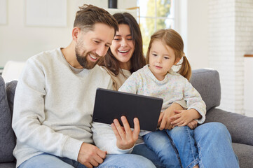 Fototapeta premium Portrait of a happy young cheerful family tablet with little cute child girl sitting on sofa at home. Mother and father enjoying weekend with daughter. Family leisure and technology concept.