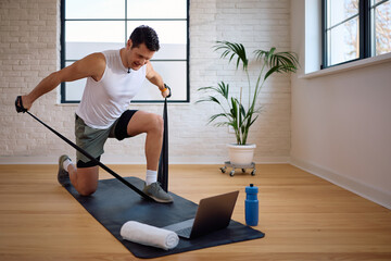 Athletic man exercising with power band while using laptop at home.